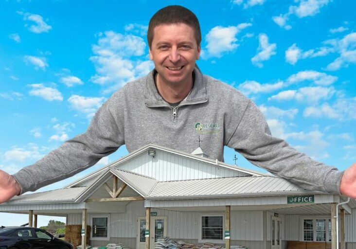 Man smiling behind a model house against a blue sky.